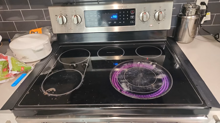 Wide view of a Samsung electric range in a residential kitchen. The bottom-right Fast Boil burner is glowing pink, and the Cooktop On indicator light is lit on the control panel, even though all knobs are in the OFF position.