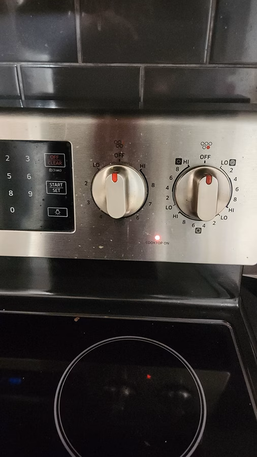 Close-up of the Samsung stove control panel showing two burner knobs both set to OFF. The Cooktop On indicator light is glowing red between them, indicating the stove believes a burner is still active.