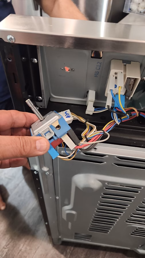 A technician holds the faulty infinite switch removed from the Samsung stove. The switch is a small electromechanical component with a metal shaft and wiring harness, shown against the open back panel of the appliance.